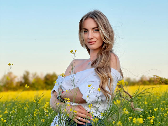 BlondyOneSex con lunghi capelli biondi ondulati, una maglietta bianca a spalle scoperte, che tiene fiori gialli, in piedi in un campo di fiori selvatici gialli, sorridente, cielo blu, giornata limpida, luce soffusa..