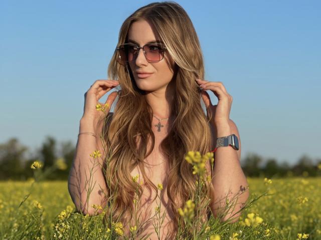 BlondyOneSex met lang, golvend blond haar, een zonnebril op, die haar haar aanraakt, een kruisbeeldketting, een blauw horloge, staand in een veld met gele wilde bloemen, een helderblauwe lucht..