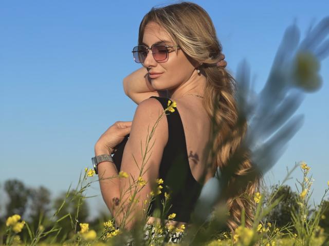 BlondyOneSex mit langen, welligen blonden Haaren, die eine Sonnenbrille und ein schwarzes Top ohne Ärmel trägt, steht in einem Feld voller gelber Blumen, blickt über ihre Schulter und zeigt ihre Brüste..