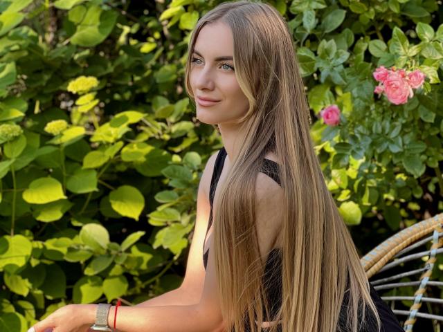 BlondyOneSex met lang, steil, blond haar, gekleed in een zwarte jurk, zittend buiten, lachend, groene ogen, rode armband, rieten stoel, roze rozen op de achtergrond..
