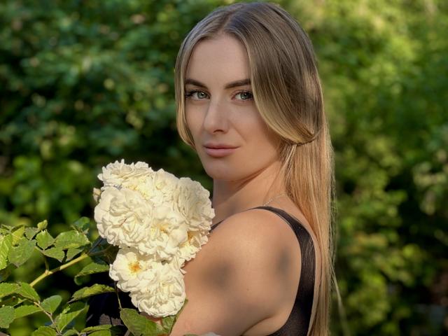 BlondyOneSex, con lunghi capelli biondi, tiene in mano rose bianche, ha un viso carino e occhi verdi, indossa una maglietta nera, in un giardino illuminato dal sole..