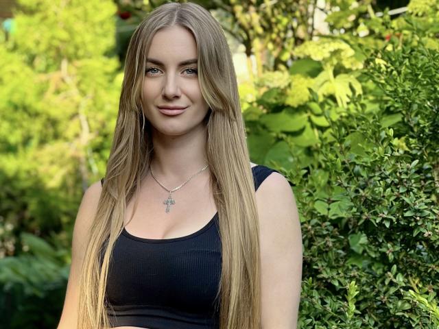 BlondyOneSex tiene el pelo rubio largo y liso, ojos azules, y lleva un top negro que deja ver sus pequeños pechos. Está sonriendo y lleva un collar de plata..