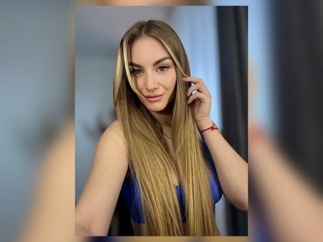 BlondyOneSex hat langes, glattes blondes Haar, einen blauen BH und ein süßes Lächeln. Sie schaut in die Kamera, wobei sie mit einer Hand ihr Haar berührt und trägt ein rotes Armband..