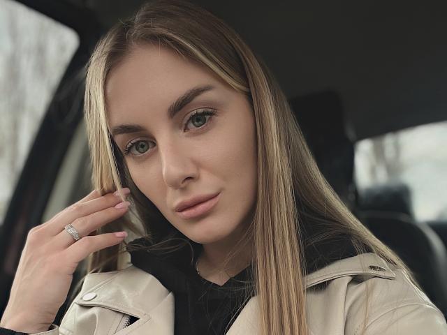 BlondyOneSex hat langes blondes Haar, grüne Augen und trägt eine beige Jacke. Sie berührt ihr Haar, zeigt einen Ring an ihrem Finger und hat ein leichtes Lächeln..