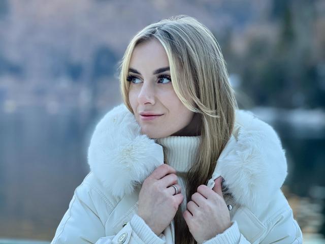 BlondyOneSex met blond haar, blauwe ogen, een witte jas met bontvoering, die een halsband vasthoudt, een lichte glimlach, een ring aan de rechterhand, een onscherpe achtergrond, koud weer, handen in de buurt van het gezicht..