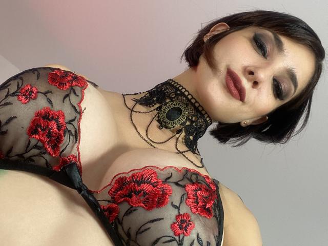 LolaGinger mit kurzen, braunen Haaren, trägt einen transparenten, schwarzen BH mit Blumenmuster, der große Brüste zeigt, mit roter Blumenstickerei, einem schwarzen Choker mit Ketten und dunklem Lippenstift, und blickt nach unten in die Kamera..