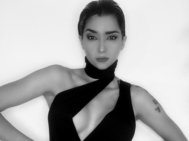 LolaGinger met glad achterover gekamd haar, gekleed in een zwarte haltertopjurk met een diepe decolleté, waarbij haar borsten en tatoeage op haar rechterarm zichtbaar zijn, op een zwart-wit foto..