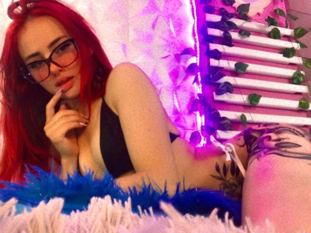 Nia Spencer, com cabelo ruivo, óculos, sutiã preto e tatuagens, deitada em um material azul e branco fofo, com o dedo nos lábios, luz neon roxa ao fundo, suporte de toalhas branco..