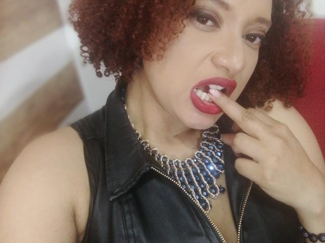 VioletaSwitch with curly red hair, black leather vest, and blue gem necklace, biting her red-lipped, white-teethed lower lip, holding her finger to her mouth.