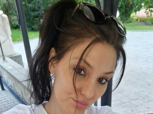 JoliDanielle has light skin, dark brown hair in a messy ponytail, black sunglasses on her head, and is pouting with pink lips. She's wearing a white shirt.