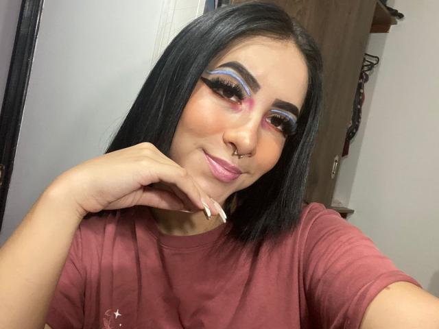 Milena Rossi mit glattem, schwarzem Haar, rosa Lidschatten, blauem Eyeliner, rosa Lippenstift, einem Nasenring und langen Nägeln, trägt ein rotes Hemd und lächelt in die Kamera..