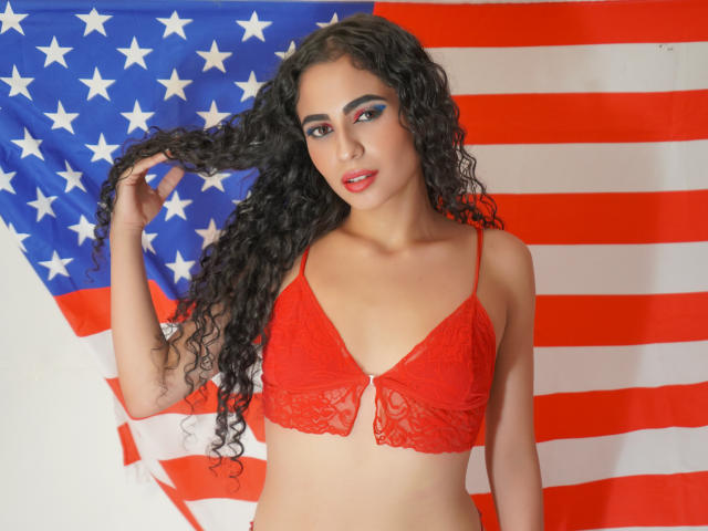 PerfectLyly, con cabello negro rizado, vistiendo un sujetador de encaje rojo, sosteniendo su cabello, de pie frente a una bandera estadounidense, tiene una mirada sexy y segura, y lleva lápiz labial rojo..