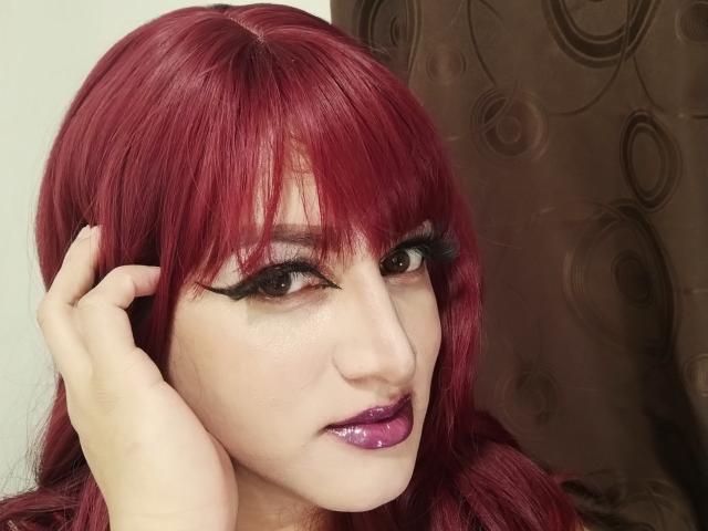 SophiaHillsxo has red hair, dark eyeliner, and glossy purple lipstick. She's touching her head with a hand, showing off her full lips and black eyelashes.