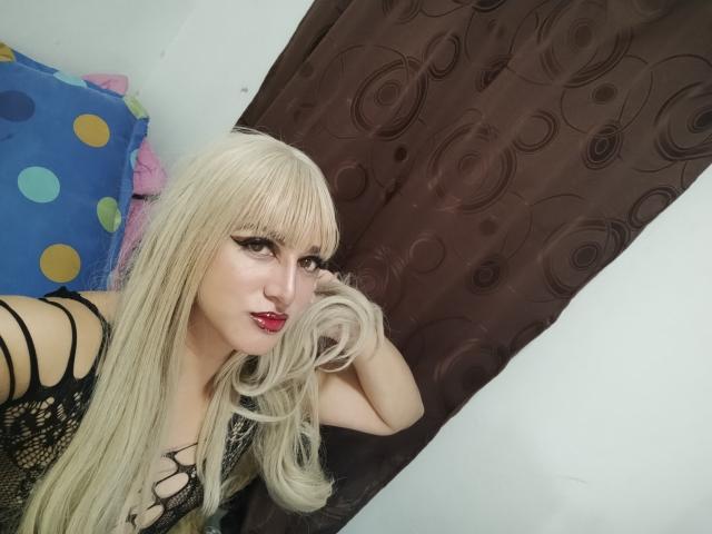SophiaHillsxo with long blonde hair, black lace top, holding her hair, red lipstick, looking at camera, brown curtain, blue polka dot pillow in background.