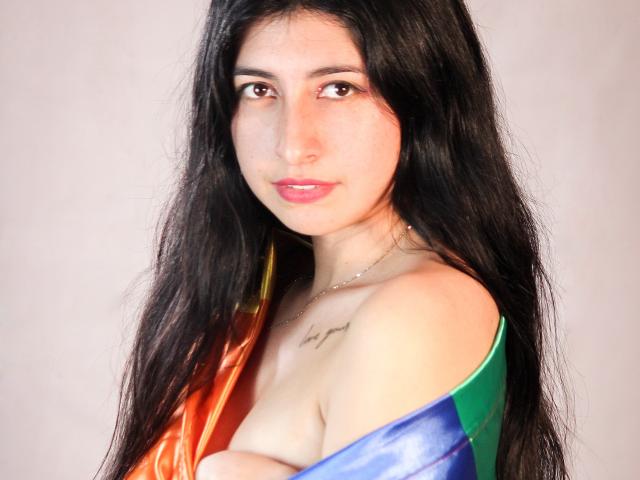EmmaXWilliams with long black hair, brown eyes, pink lips, and a small tattoo on her right shoulder, wearing a rainbow-colored dress, showing her bare shoulder and chest.