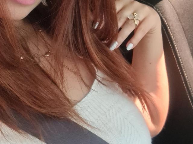 De borst van GiovannaSweet, met een witte gebreide top, bruin haar, een grijze veiligheidsgordel, een gouden kruisring, witte nagels, roze lippen en een bruine jas..