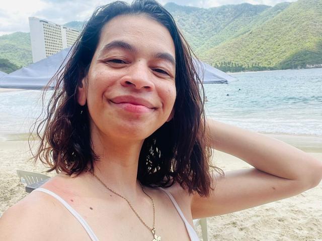 AmberGray69, smiling with wet brown hair, small nose, and slight freckles, wearing a white tank top, hand behind head, necklace, beach, mountains in background, water and sand.