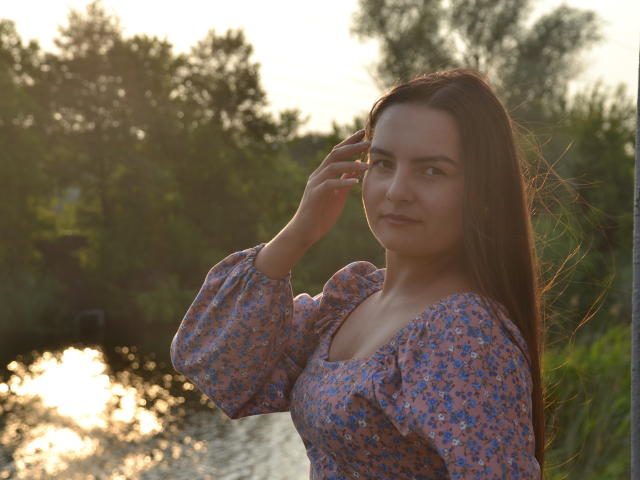 JasmineAsha, con cabello largo y castaño, vestida con un vestido floral, de pie junto a un río al atardecer, tocándose el cabello, mirando ligeramente hacia un lado con una expresión tranquila..