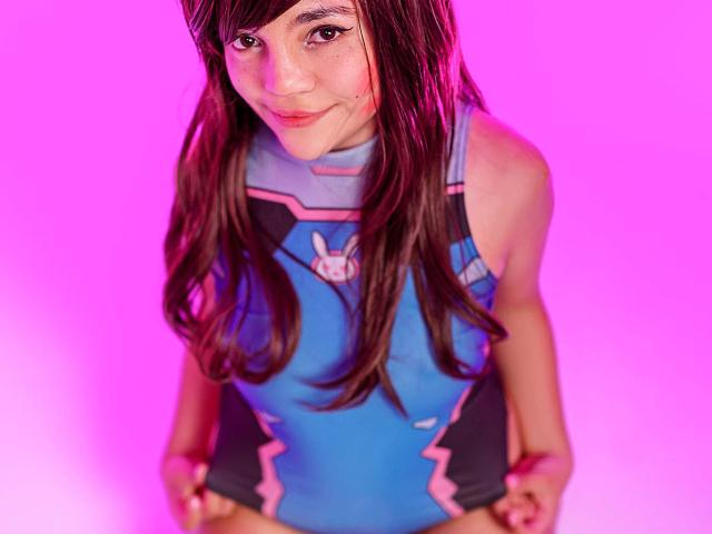DreamySweetLucy, vestindo um cosplay azul e rosa da D.Va, com cabelos castanhos longos, sardas e um sorriso adorável, exibindo seus pequenos seios, em um fundo iluminado em rosa..