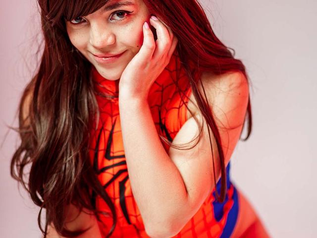DreamySweetLucy, avec de longs cheveux bruns, portant un costume rouge de Spider-Man, touchant sa joue avec un léger sourire, montrant ses seins dans la tenue ajustée et colorée..
