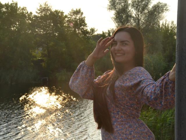 JasmineAsha with long brown hair, smiling, wearing a blue floral dress, standing by a sunlit river, raising her right hand, sunlight reflecting off the water.