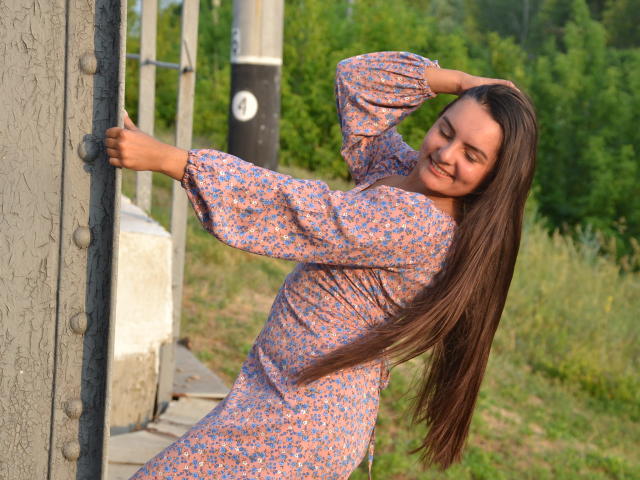 JasmineAsha with long brown hair, wearing a floral dress, leaning against a metal pole, smiling, eyes closed, right hand touching her head, left arm pointing, outdoors.
