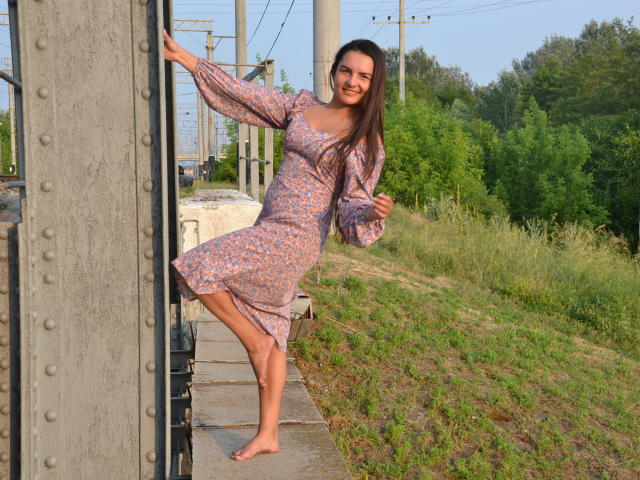 JasmineAsha, pieds nus, dans une robe fleurie, debout sur les voies ferrées, appuyée contre un poteau métallique, souriante, avec de longs cheveux bruns, et une jolie silhouette, entourée de verdure..
