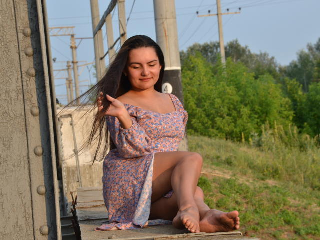 JasmineAsha with long brown hair, wearing a purple floral dress, sitting barefoot on a train, touching her hair, smiling, showing her tits and ass, in a sunny outdoor setting.