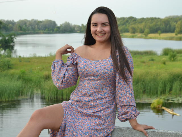 JasmineAsha with long brown hair, smiling, wearing a pink floral off-shoulder dress, sitting on a stone ledge, touching her hair, showing her bare thigh, by a calm waterway.