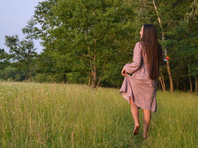 JasmineAsha with long brown hair, wearing a floral dress, stands in a grassy field, holding a red flower, her ass slightly visible, facing away, in brown sandals.