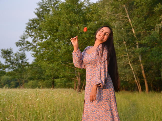 JasmineAsha with long dark hair, wearing a floral dress, holding a red flower, smiling, standing in a grassy field, evening light, her ass slightly visible in the dress.