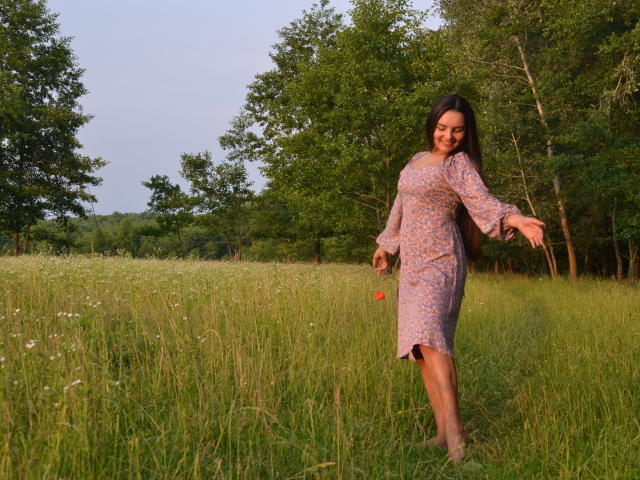 JasmineAsha with long brown hair, wearing a pink floral dress, stands in a grassy field, arms out, smiling, legs slightly apart, showing her ass in the sunlight.