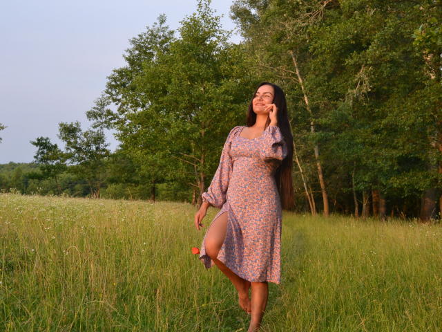 JasmineAsha stands in a grassy field, wearing a pink floral dress, lifting her right leg, smiling, with long brown hair, touching her chin, in a sunny forest.