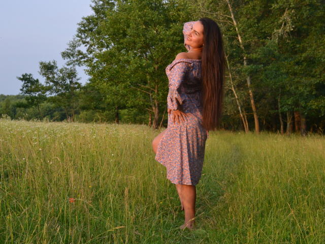 JasmineAsha with long brown hair in a pink floral dress, standing barefoot in a grassy field, looking up with hands on belly, trees in background, sun setting.