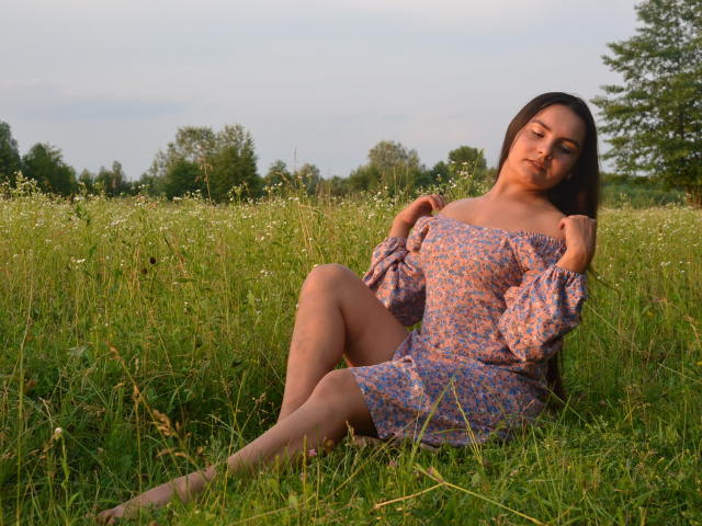 JasmineAsha, with bare tits and ass, sits in grass, wearing an off-shoulder floral dress, hands on dress, legs crossed, brown hair, soft smile, evening light, green field.