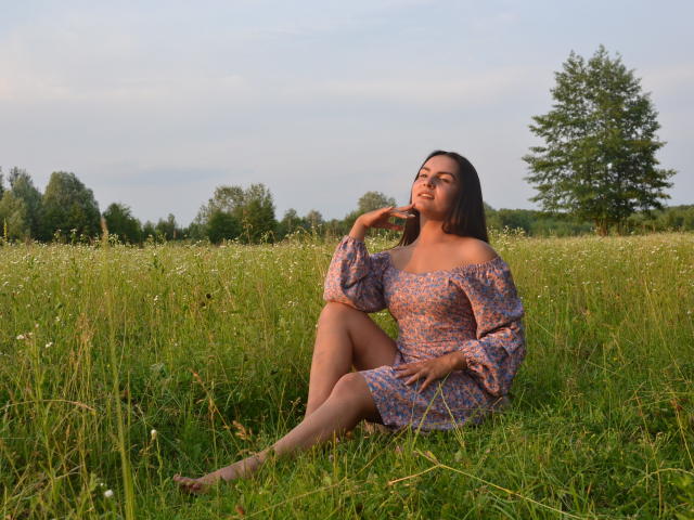 JasmineAsha sitting in a grassy field, wearing a purple floral off-shoulder dress, her hand near her face, looking up, with a tall tree in the background.