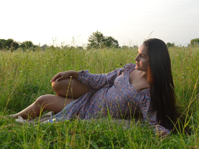 JasmineAsha with long brown hair, lying in grass, wearing a floral dress, showing her tits, legs up, right arm resting on knee, looking sideways, sunset light, calm expression.