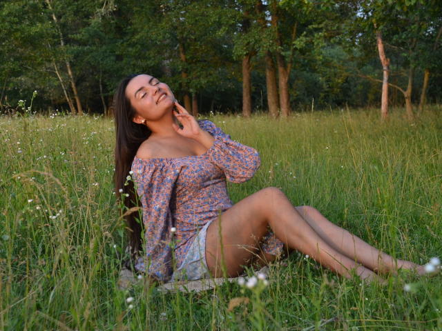 JasmineAsha with long black hair, smiling, eyes closed, off-shoulder floral dress, sitting in grass, hand on neck, legs bent, sunlit, green field with white flowers.