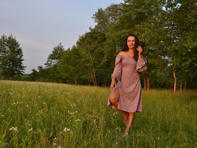 JasmineAsha with long dark hair, wearing a purple floral dress, lifting one leg, standing in a grassy field with white flowers, trees in the background, smiling at the camera.
