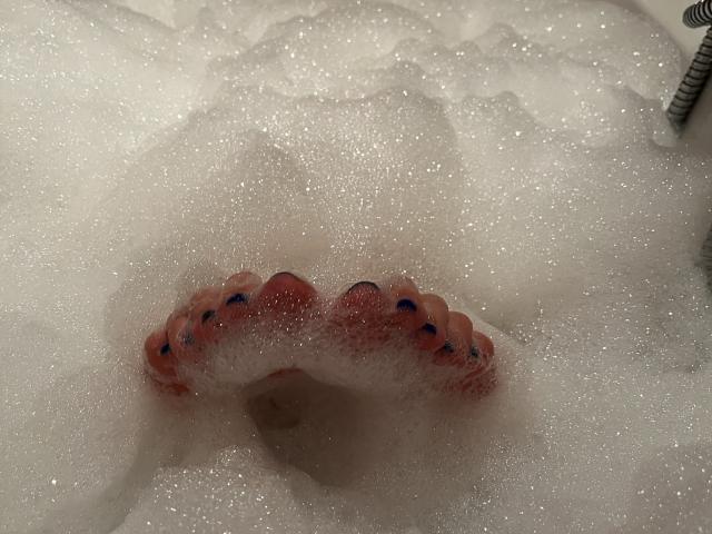 MelanieFrancaiseDuo's pink toes with blue nail polish are partially visible through a thick, foamy bubble bath. The toes are slightly curled and covered in white bubbles.