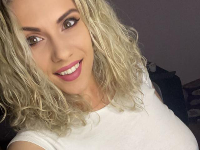 LucilleXSugar tem cabelo loiro e encaracolado, olhos castanhos e um sorriso com batom rosa. Ela está vestindo uma blusa branca, que mostra seus seios, e tem um rosto bonito e sexy..
