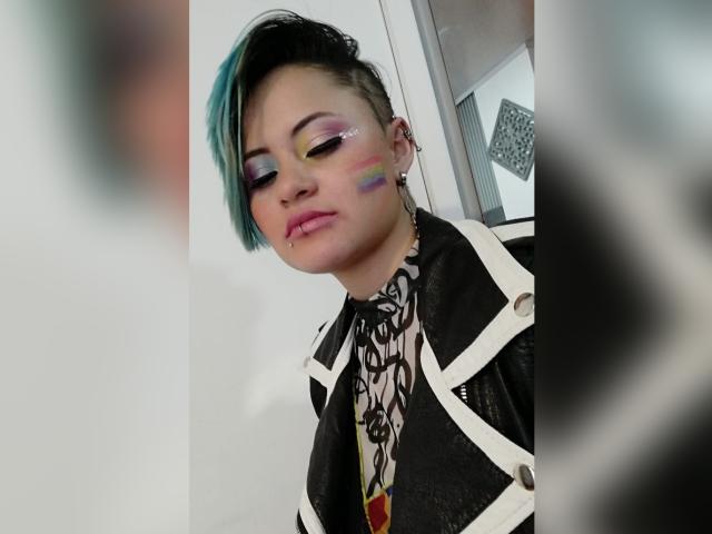 CassieJones with short black and green hair, colorful eyeshadow, rainbow cheek, nose ring, lip piercing, black and white patterned jacket, and a confident, slightly bored expression.
