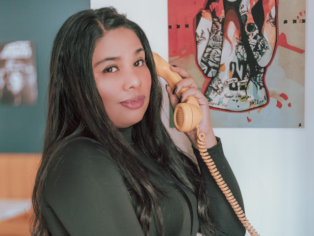TriniShely, con lunghi capelli neri, indossa un maglione nero a collo alto, tiene un telefono beige con il filo all'orecchio e guarda la fotocamera con le labbra rosa leggermente dischiuse..
