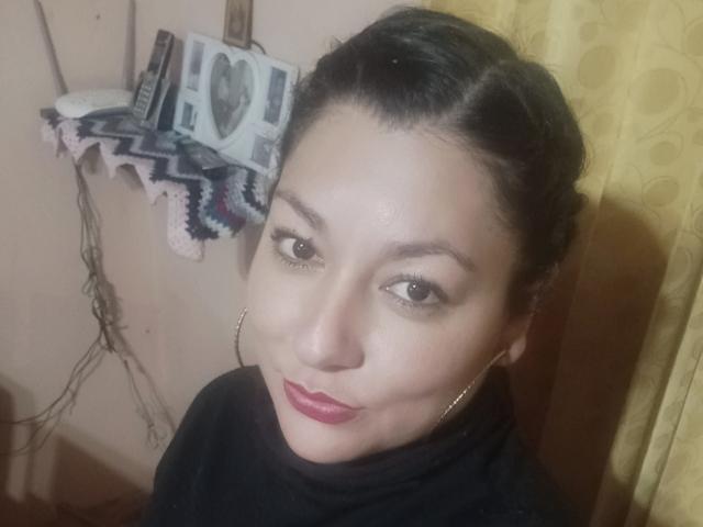 SakotoDika has medium skin, dark hair in a braid, brown eyes, red lipstick, gold hoop earrings, and wears a black shirt. Her small nose and smile are visible.
