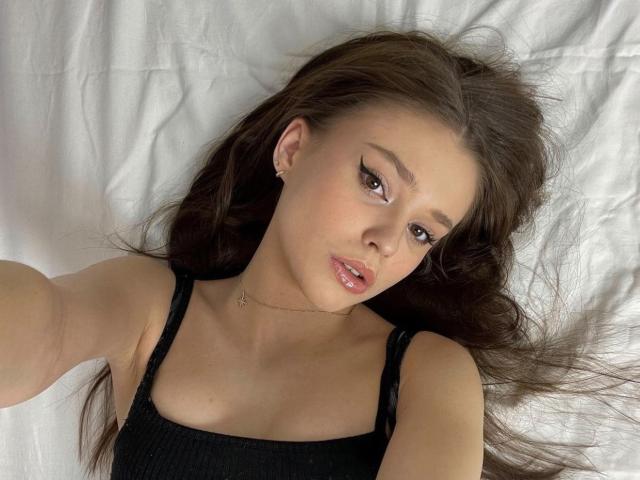 OliviyaBackers has long brown hair, big brown eyes, and shiny pink lips, wearing a black tank top, lying on a white bed, looking up at the camera.