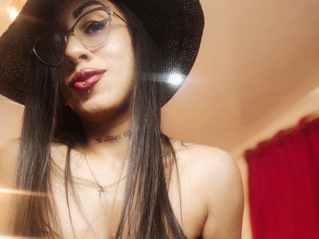 LunaCandy, con cabello negro largo, usando gafas, lápiz labial rojo, un sombrero negro con brillantina, un collar y un tatuaje visible, mirando a la cámara, con reflejos de luz en el rostro, y una cortina roja de fondo..