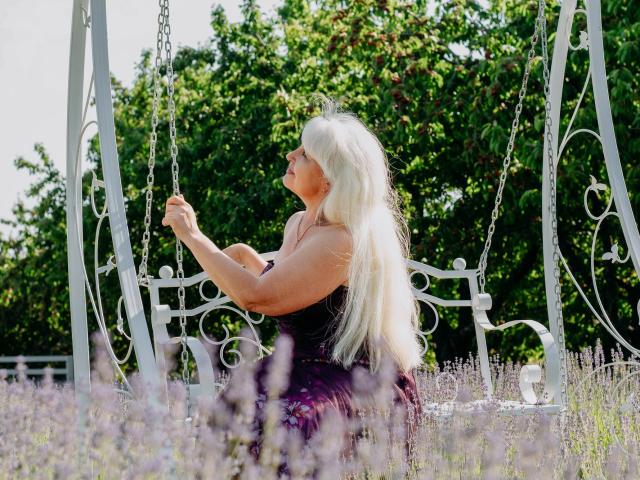 KamaliaSim with long, platinum blonde hair, sitting on a white, ornate swing, holding chains, wearing a black, strapless top, and a purple floral skirt, in a sunlit field.