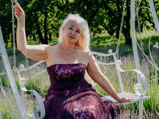 KamaliaSim, an older woman with white hair, wearing a strapless purple floral dress, sits on a white metal swing holding a chain. Her tits are small and she's smiling.