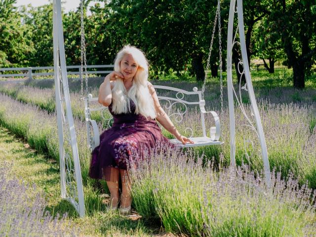 KamaliaSim, com cabelos brancos longos, vestido floral roxo, sentada em um balanço branco ornamentado em um campo de lavanda, com a mão no rosto, árvores verdes ao fundo, dia ensolarado, relaxada, fofa..