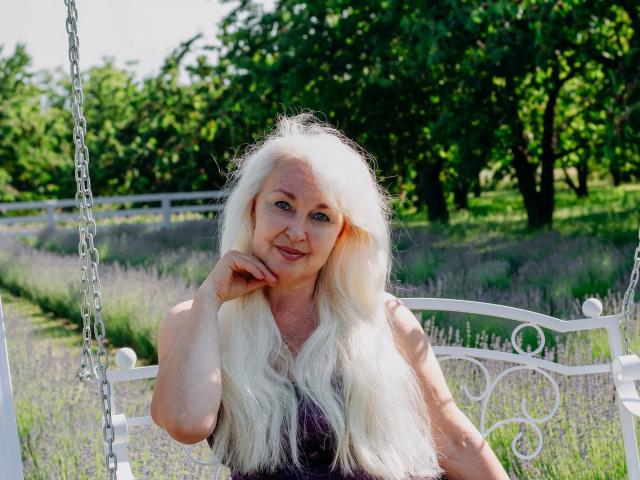 KamaliaSim with long, white, wavy hair, smiling, has a hand on her chin, wearing a dark top, sitting on a white, metal swing in a lavender field.